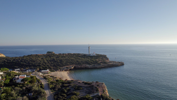 Aerial view of the lighthouse at Ponta do Altar featuring rugged cliffs coastal vegetation and the calm Atlantic Ocean under a clear sky in Ferragudo Faro District Portugal.