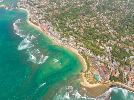 Aerial view of the turquoise ocean waves crashing against the sandy beach and coastal buildings surrounded by lush tropical greenery in Hikkaduwa Southern Province Sri Lanka. Aerial view of the turquoise ocean waves crashing against the sandy beach and coastal buildings surrounded by lush tropical greenery in Hikkaduwa Southern Province Sri Lanka.