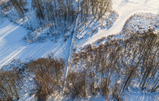 Aerial view of a narrow wooden bridge crossing a snow covered landscape surrounded by bare trees and long shadows in Lithuania. Aerial view of a narrow wooden bridge crossing a snow covered landscape surrounded by bare trees and long shadows in Lithuania.