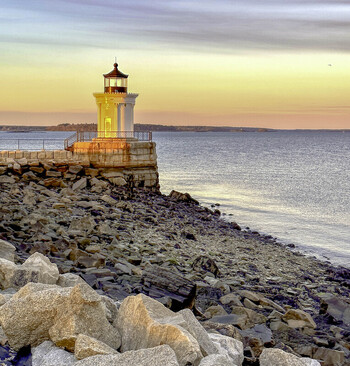 Aerial view of Portland Breakwater Lighthouse also known as Bug Light situated on a rocky shoreline at sunset with calm water and golden light in Portland Maine United States.