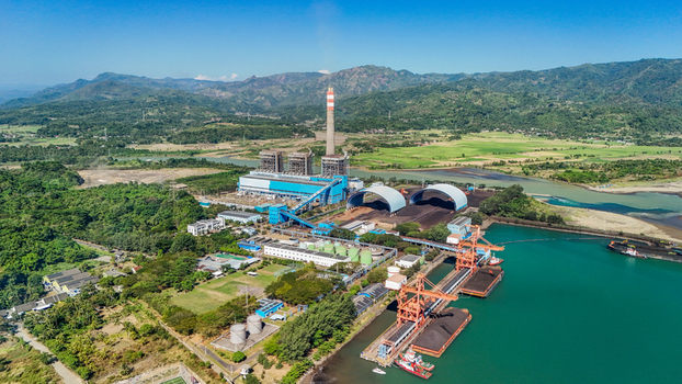 Aerial view of a coal fired power plant situated along a riverbank with industrial infrastructure storage domes and cargo ships docked in Indonesia.