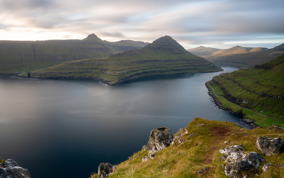 View of the dramatic fjord landscape featuring lush green mountains calm dark waters and a rocky foreground under a soft cloudy sky in the Faroe Islands. View of the dramatic fjord landscape featuring lush green mountains calm dark waters and a rocky foreground under a soft cloudy sky in the Faroe Islands.