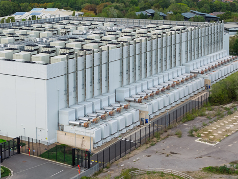Aerial view of a large data center facility with rows of cooling units and ventilation systems on the roof surrounded by a security fence and paved grounds in the United Kingdom.