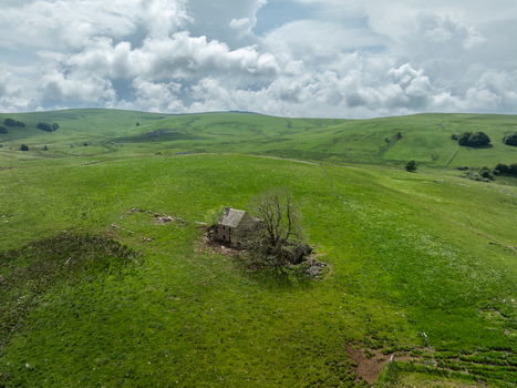 Aerial view of a solitary stone building nestled amidst rolling green hills under a cloudy sky Nasbinals Occitanie France. Aerial view of a solitary stone building nestled amidst rolling green hills under a cloudy sky Nasbinals Occitanie France.