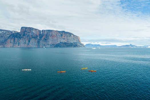 Aerial view of kayakers paddling in the deep blue waters of the bay with the massive Uummannaq mountain rising in the background in Uummannaq Kitaa Greenland. Aerial view of kayakers paddling in the deep blue waters of the bay with the massive Uummannaq mountain rising in the background in Uummannaq Kitaa Greenland.
