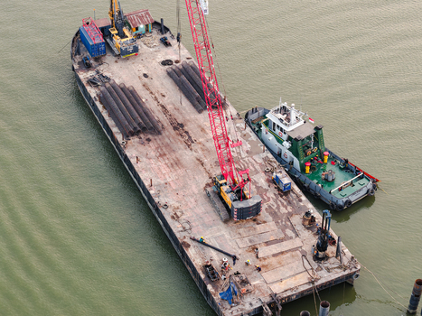 Aerial view of a construction barge on the Mahakam River with a red crane steel pipes and a tugboat alongside in Samarinda East Kalimantan Indonesia. Aerial view of a construction barge on the Mahakam River with a red crane steel pipes and a tugboat alongside in Samarinda East Kalimantan Indonesia.