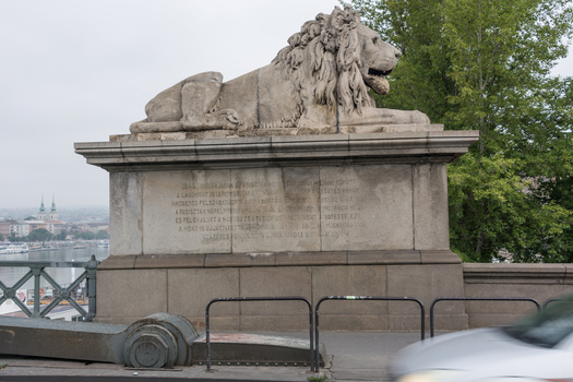 View of the stone lion statue on the Szechenyi Chain Bridge with an inscription on the pedestal and a blurred car passing by in Budapest Hungary.