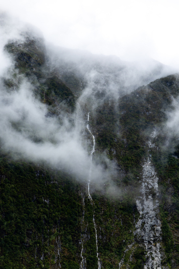 View of a misty mountain face shrouded in low hanging clouds with a thin waterfall cascading down the dark forested slopes Milford Sound Southland Region New Zealand.
