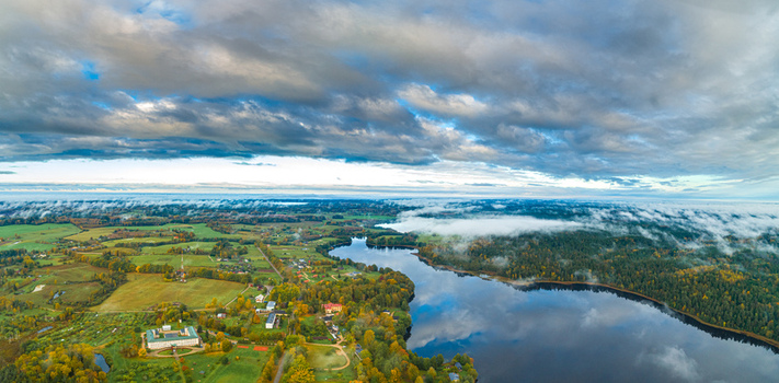 Aerial view of a serene lake surrounded by lush autumn forests green fields and scattered buildings under a dramatic cloudy sky in Latvia. Aerial view of a serene lake surrounded by lush autumn forests green fields and scattered buildings under a dramatic cloudy sky in Latvia.