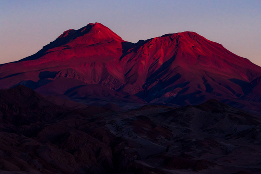 Aerial view of red mountain peaks illuminated by the setting sun above dark desert hills in Toconao Antofagasta Chile. Aerial view of red mountain peaks illuminated by the setting sun above dark desert hills in Toconao Antofagasta Chile.