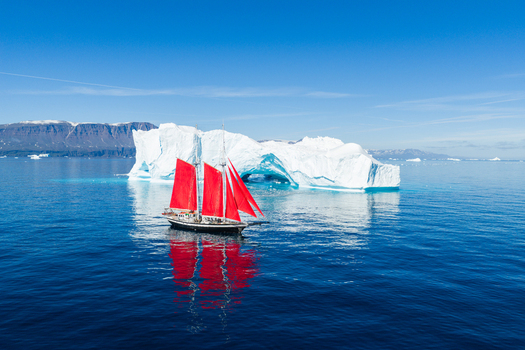 Aerial view of a sailboat with vibrant red sails navigating the deep blue waters near a massive white iceberg with a natural arch set against a mountainous coastline in Greenland. Aerial view of a sailboat with vibrant red sails navigating the deep blue waters near a massive white iceberg with a natural arch set against a mountainous coastline in Greenland.