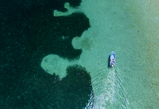 Aerial view of a small blue boat with people cruising through turquoise and dark green waters over coral reefs in Guanaja Bay Islands Department Honduras. Aerial view of a small blue boat with people cruising through turquoise and dark green waters over coral reefs in Guanaja Bay Islands Department Honduras.