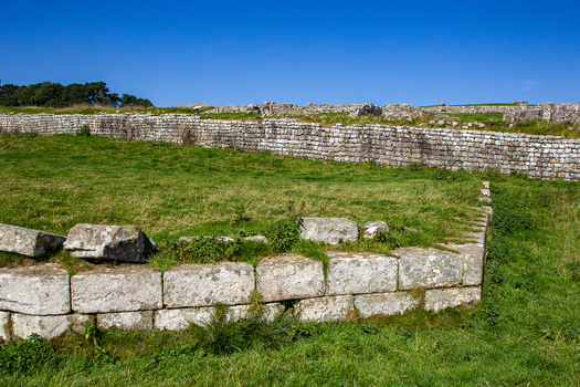 View of the ancient stone walls and excavated ruins of Vindolanda Roman Fort set against green grassy fields and a clear blue sky in Hexham England United Kingdom. View of the ancient stone walls and excavated ruins of Vindolanda Roman Fort set against green grassy fields and a clear blue sky in Hexham England United Kingdom.