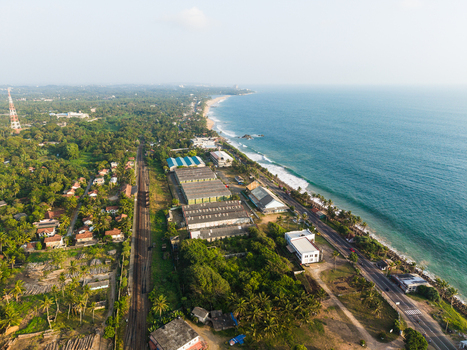 Aerial view of the tropical coastline featuring a railway track industrial warehouses and lush greenery along the ocean in Galle Southern Province Sri Lanka.