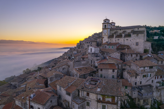 Aerial view of the historic hilltop town of Artena with its stone buildings and terracotta rooftops rising above a sea of fog during a golden sunrise in Artena Lazio Italy.
