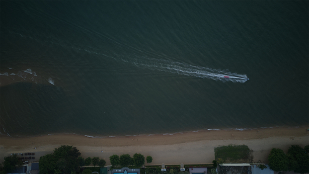 Aerial view of Jomtien Beach with a boat leaving a long white wake in the dark sea along the sandy coastline and lush green trees in Chon Buri Thailand.