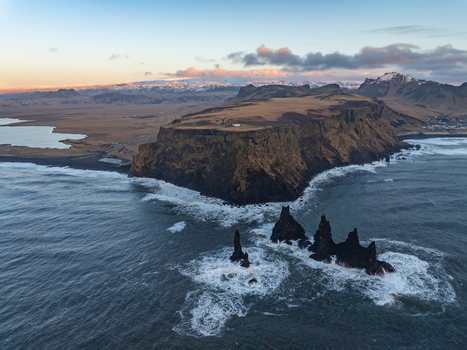 Aerial view of the Dyrholaey promontory with its dramatic sea stacks crashing waves and rugged coastline under a soft golden hour sky in Myrdalshreppur Iceland. Aerial view of the Dyrholaey promontory with its dramatic sea stacks crashing waves and rugged coastline under a soft golden hour sky in Myrdalshreppur Iceland.