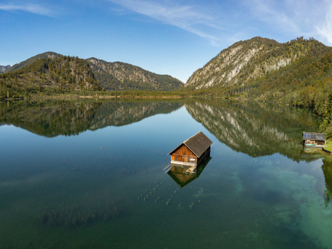 Aerial view of a wooden boathouse floating on the calm clear waters of Attensee surrounded by forested mountains under a bright blue sky in Rottenbach Bayern Germany. Aerial view of a wooden boathouse floating on the calm clear waters of Attensee surrounded by forested mountains under a bright blue sky in Rottenbach Bayern Germany.