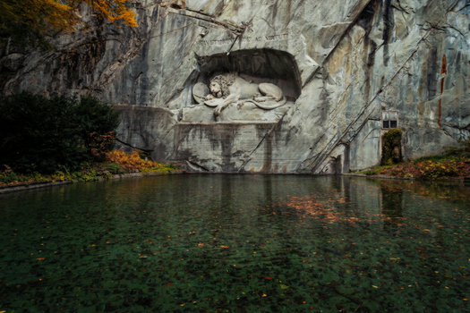 View of the Lion Monument carved into a sandstone rock face above a calm pond with autumn leaves in Lucerne Lucerne Switzerland. View of the Lion Monument carved into a sandstone rock face above a calm pond with autumn leaves in Lucerne Lucerne Switzerland.