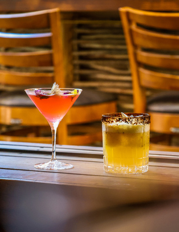 View of a pink martini with flower petals and a yellow rocks cocktail with a dark rim on a wooden table at Rubas Bakery and Bistro in San Jose del Cabo Baja California Sur Mexico.
