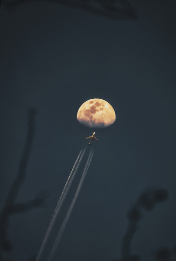 View of a commercial airplane flying in front of a bright moon with long contrails trailing behind in the night sky over Shanghai Shanghai China.