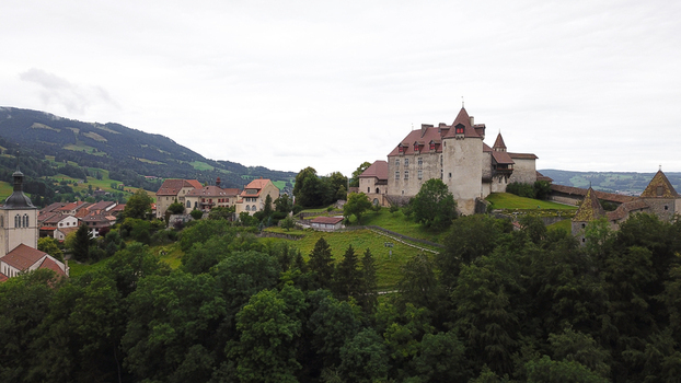 Aerial view of Chateau de Gruyeres perched on a lush green hill overlooking the medieval village and rolling alpine foothills under a cloudy sky Gruyeres Fribourg Switzerland.