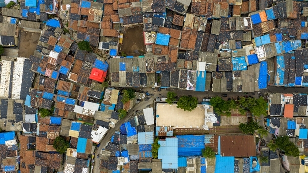 Aerial view of the dense Dharavi Slums featuring a patchwork of corrugated metal and blue plastic roofs narrow alleyways and a white roofed building in Mumbai Maharashtra India. Aerial view of the dense Dharavi Slums featuring a patchwork of corrugated metal and blue plastic roofs narrow alleyways and a white roofed building in Mumbai Maharashtra India.