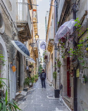 View of a figure walking down a narrow alleyway adorned with plants umbrellas and architectural details evoking a sense of depth and charm Ortigia Sicily Italy.