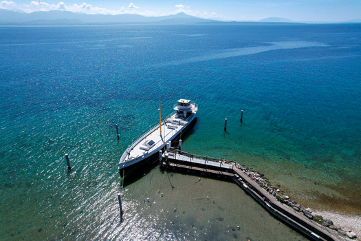 Aerial view of a white passenger boat docked at a wooden pier on the turquoise waters of Lake Geneva with mountains in the distance Perroy Vaud Switzerland. Aerial view of a white passenger boat docked at a wooden pier on the turquoise waters of Lake Geneva with mountains in the distance Perroy Vaud Switzerland.