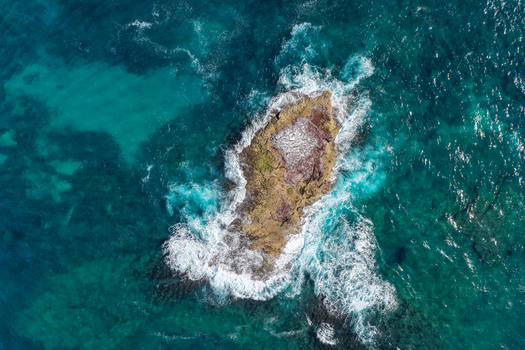 Aerial view of a rocky outcrop off the coast of North Stradbroke Island surrounded by turquoise ocean waves crashing against the stone in Redland City Queensland Australia. Aerial view of a rocky outcrop off the coast of North Stradbroke Island surrounded by turquoise ocean waves crashing against the stone in Redland City Queensland Australia.