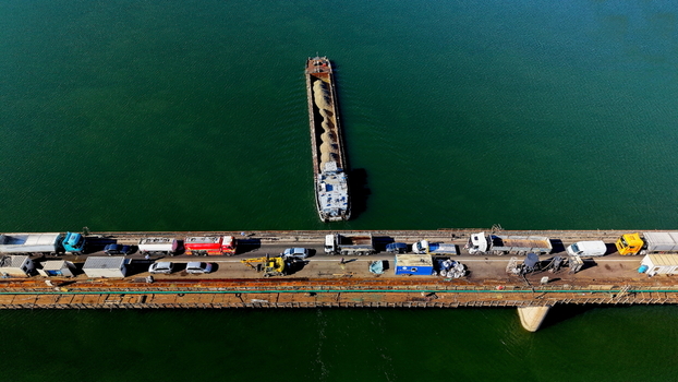 Aerial view of a barge transporting sand on the Danube river passing by a pier with parked trucks and construction equipment in Smederevo Podunavlje District Serbia.