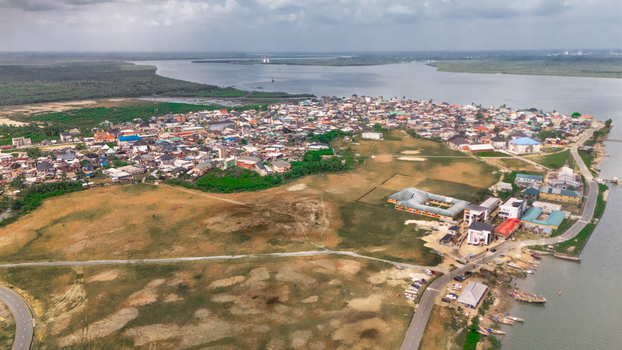 Aerial view of a coastal town with dense residential buildings winding roads and boats docked along the riverbank under a cloudy sky in Rivers State Nigeria.