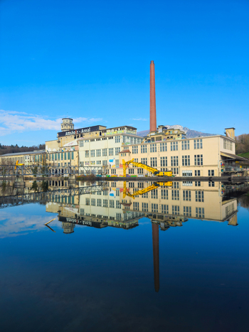 View of the Former Cellulose Factory Attisholz Areal with its tall chimney and industrial buildings reflecting in the calm river water under a clear blue sky Solothurn Solothurn Switzerland. View of the Former Cellulose Factory Attisholz Areal with its tall chimney and industrial buildings reflecting in the calm river water under a clear blue sky Solothurn Solothurn Switzerland.