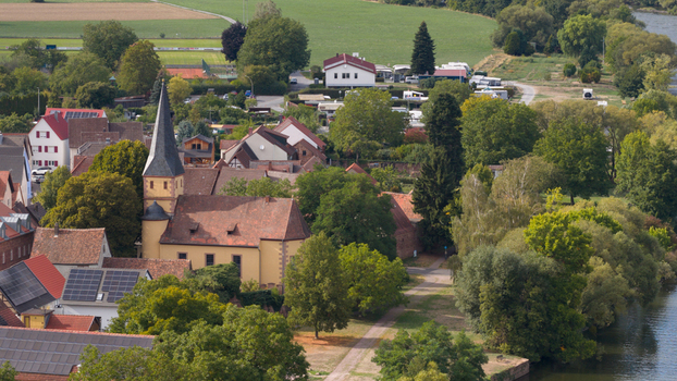 Aerial view of a historic church with a tall steeple surrounded by traditional red tiled rooftops lush green trees and a river in Kleinheubach Bavaria Germany.