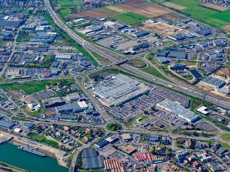 Aerial view of sprawling highway interchanges and a large shopping center with vast parking lots contrasting against the verdant fields and urban sprawl Sausheim Grand Est France.