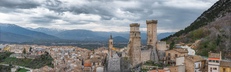 Aerial view of Caldora Castle and the historic stone buildings of the village under a dramatic cloudy sky with mountains in the background Pacentro Abruzzo Italy. Aerial view of Caldora Castle and the historic stone buildings of the village under a dramatic cloudy sky with mountains in the background Pacentro Abruzzo Italy.
