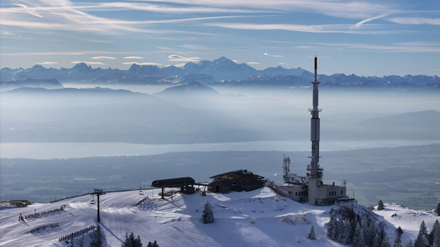 Aerial view of a snow covered mountain peak with a communications tower piercing through the crisp air overlooking the hazy valley and distant Alps Gex Auvergne Rhône Alpes France. Aerial view of a snow covered mountain peak with a communications tower piercing through the crisp air overlooking the hazy valley and distant Alps Gex Auvergne Rhône Alpes France.