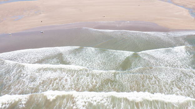 Aerial view of Bantham Beach with rolling waves and white foam on the sandy shore as surfers walk along the water in Kingsbridge Devon United Kingdom. Aerial view of Bantham Beach with rolling waves and white foam on the sandy shore as surfers walk along the water in Kingsbridge Devon United Kingdom.