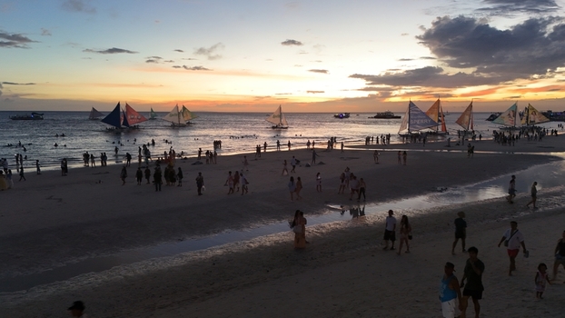 Malay Philippines 02 May 2026 Aerial view of Boracay Island at sunset with many tourists on the white sand beach and traditional paraw sailboats on the calm sea under a golden sky.
