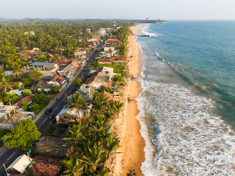 Aerial view of the sandy coastline and turquoise ocean waves alongside a coastal road lined with palm trees and houses in Galle Southern Province Sri Lanka.