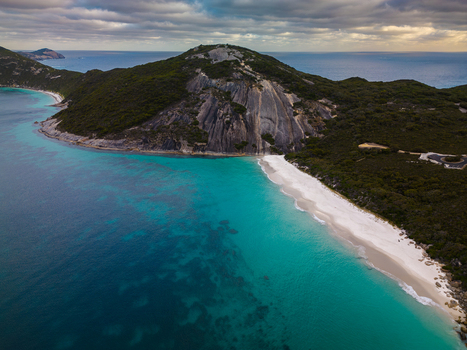 Aerial view of Misery Beach featuring white sand and turquoise waters next to a massive granite cliff under a cloudy sky in Torndirrup Western Australia Australia.