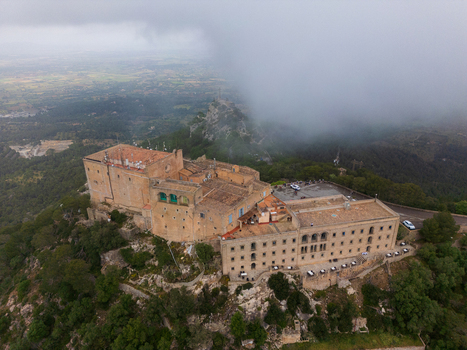 Aerial view of the Sanctuary of Sant Salvador with historic stone architecture on a hilltop surrounded by lush green trees under a misty sky in Felanitx Illes Balears Spain. Aerial view of the Sanctuary of Sant Salvador with historic stone architecture on a hilltop surrounded by lush green trees under a misty sky in Felanitx Illes Balears Spain.