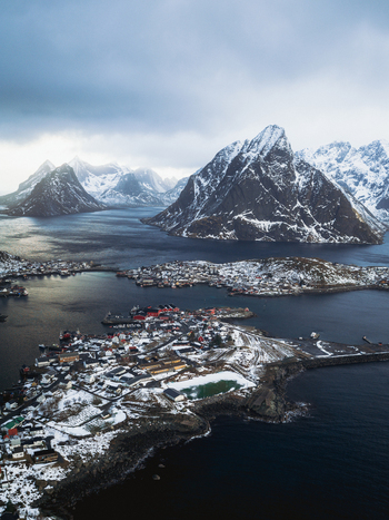 Aerial view of Reinebringen s dramatic fjords and snow dusted peaks meet the vibrant red roofed village nestled along the rugged coastline Reinebringen Nordland Norway.