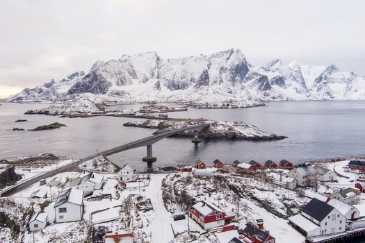 Aerial view of the snow covered mountains and traditional red wooden houses of Lofoten connected by a bridge in the winter landscape of Nordland Norway.