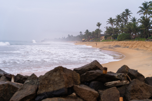 View of a sandy tropical beach with crashing waves palm trees lining the shore and dark rocks in the foreground under an overcast sky in Galle Southern Province Sri Lanka.