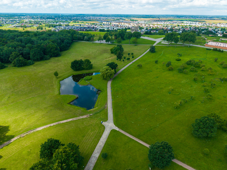 Aerial view of the IX fortas memorial park featuring a small pond walking paths and lush green lawns with a distant cityscape under a cloudy sky in Kaunas Kaunas County Lithuania.