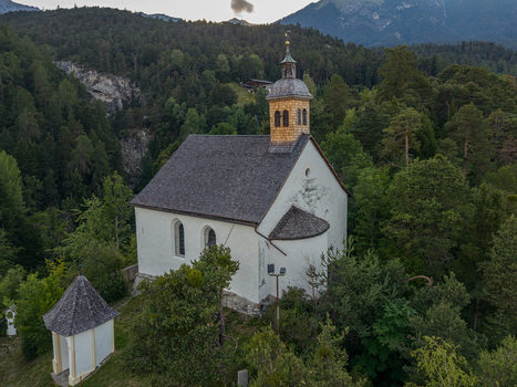 Aerial view of St. Laurentius Church with its distinctive wooden steeple nestled among dense green forests and the Austrian Alps in Imst Tyrol Austria.
