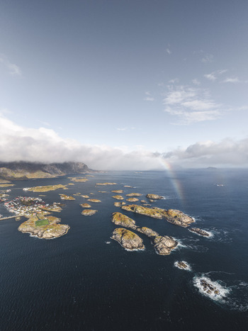 Aerial view of the Lofoten archipelago featuring rocky islands calm dark blue sea and a faint rainbow under a cloudy sky in Leknes Nordland Norway. Aerial view of the Lofoten archipelago featuring rocky islands calm dark blue sea and a faint rainbow under a cloudy sky in Leknes Nordland Norway.