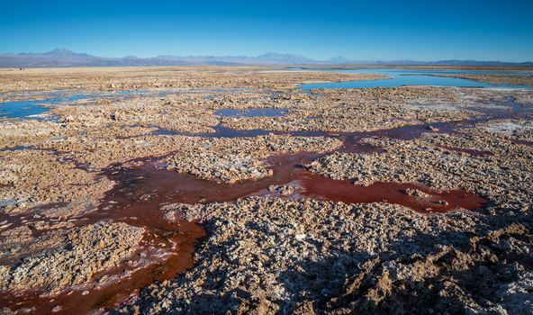 View of Laguna Chaxa salt flats with reddish mineral rich water rugged salt crust and distant mountains under a clear blue sky in San Pedro de Atacama Antofagasta Chile.