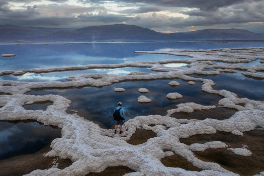 Aerial view of a lone figure exploring the surreal brilliant white salt formations contrasting with the deep blue waters of the Dead Sea under a dramatic sky Dead Sea Israel. Aerial view of a lone figure exploring the surreal brilliant white salt formations contrasting with the deep blue waters of the Dead Sea under a dramatic sky Dead Sea Israel.